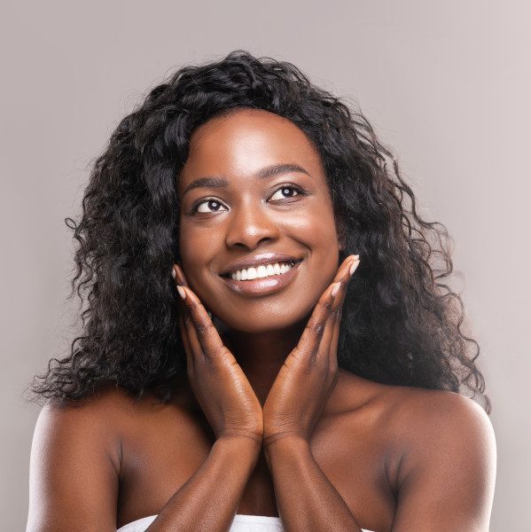 Young african woman touching her smooth cheeks, applying moisturizer cream on her pretty face, grey studio background